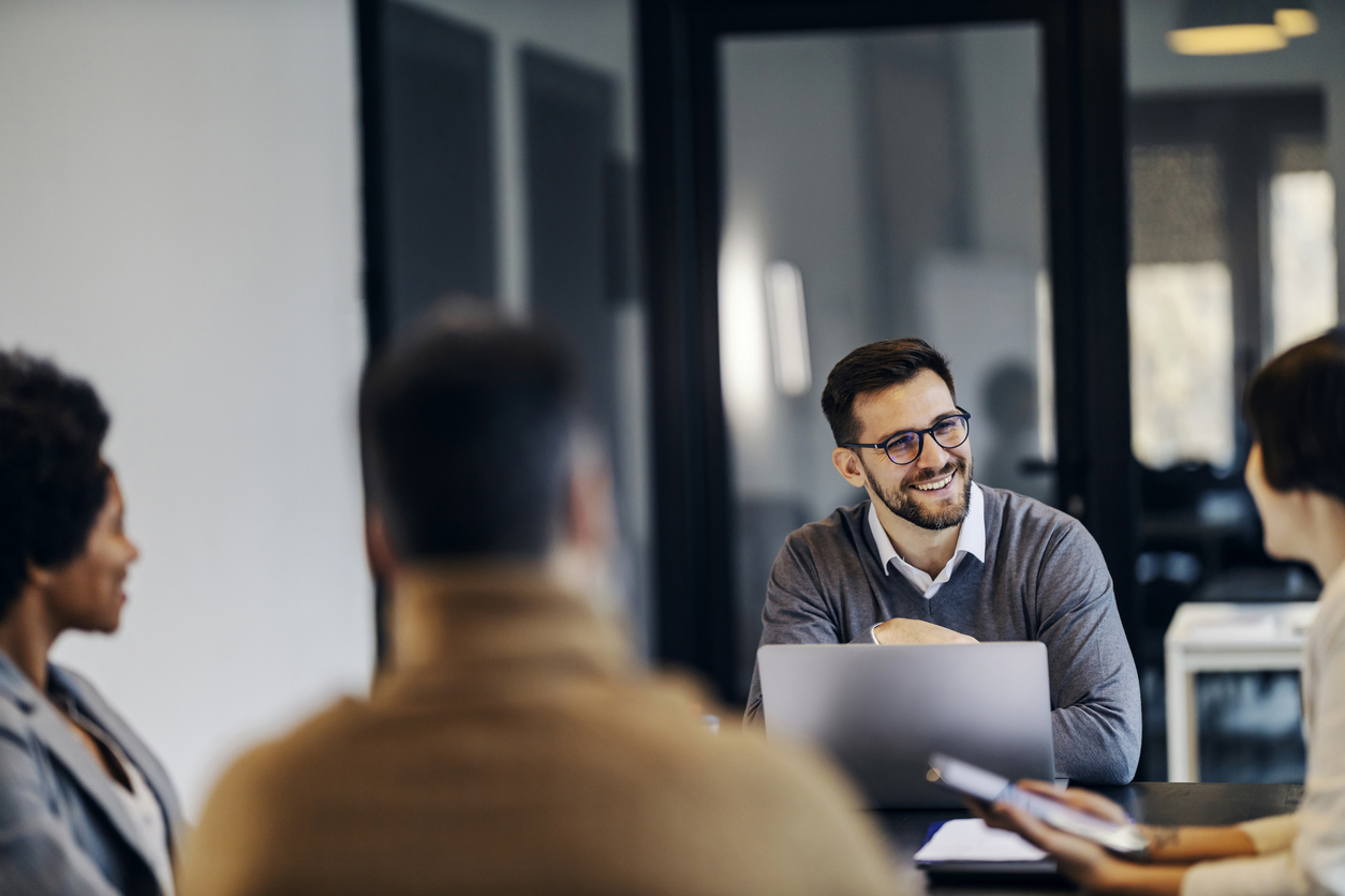 A happy businessman is sitting in the co working space with colleagues and having briefing.